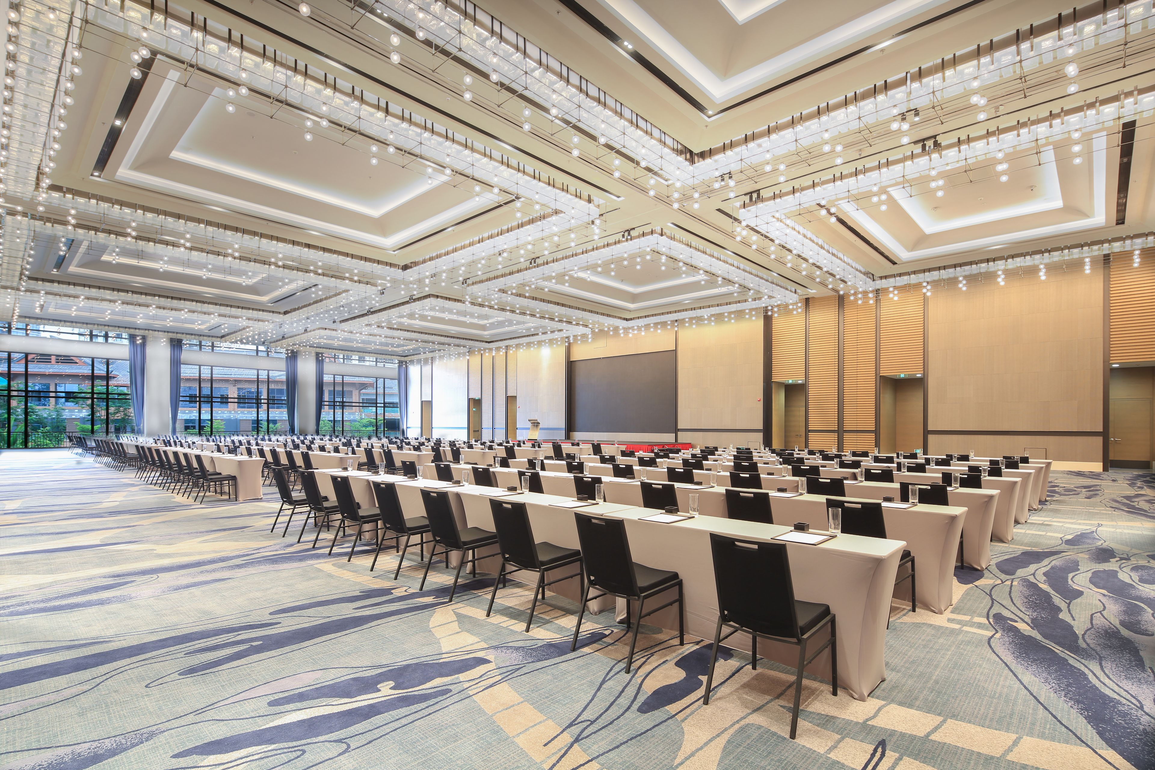 A conference room with rows of tables and chairs, a large screen, and a high ceiling with lighting fixtures.