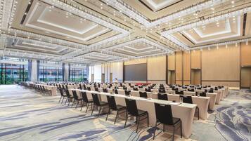 A conference room with rows of tables and chairs, a large screen, and a high ceiling with lighting fixtures.