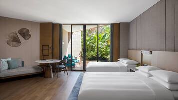 A modern hotel room with a large bed, a sofa, a small round table, and a view of greenery through the glass door.