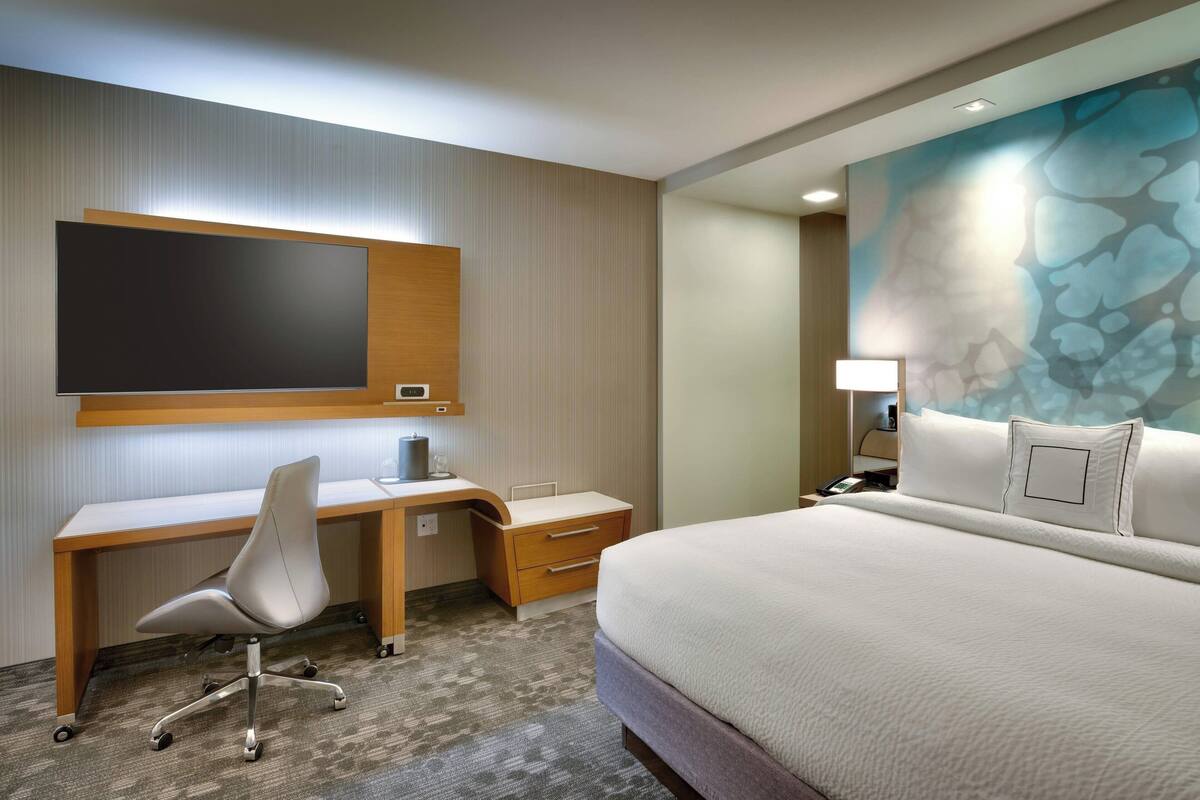 Habitación de hotel con cama, escritorio con silla, televisor de pantalla plana, mesita de noche y un cuadro abstracto grande en la pared.