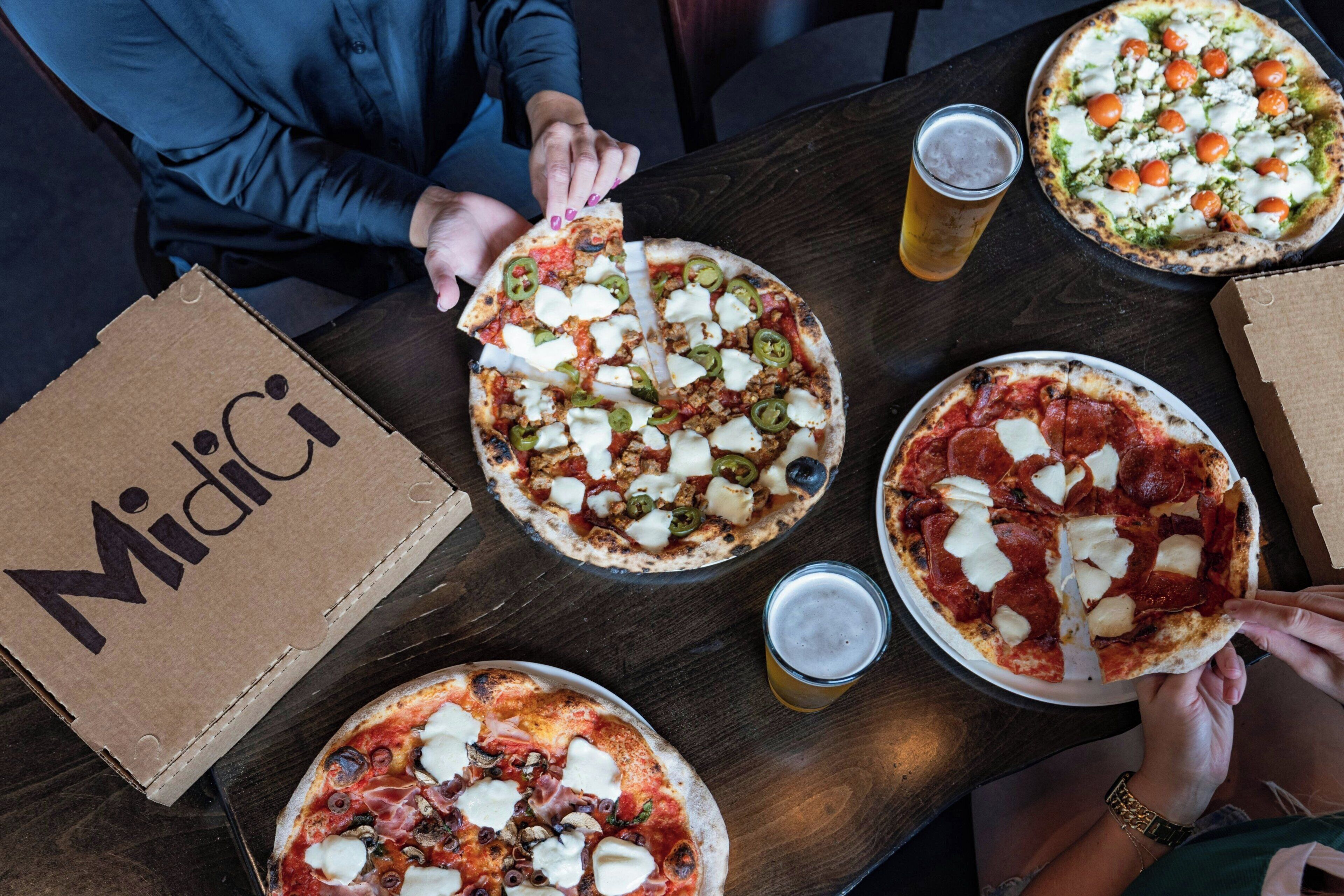 Una mesa con tres pizzas, dos bebidas y una caja con la etiqueta 'Midici'.