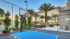 Una cancha de baloncesto al aire libre con piso azul, rodeada por una cerca, palmeras y un edificio con zona de piscina al fondo.