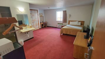 Quarto de hotel com tapete vermelho, uma cama, escrivaninha com cadeira, espelho, cabideiro e janela com cortinas.