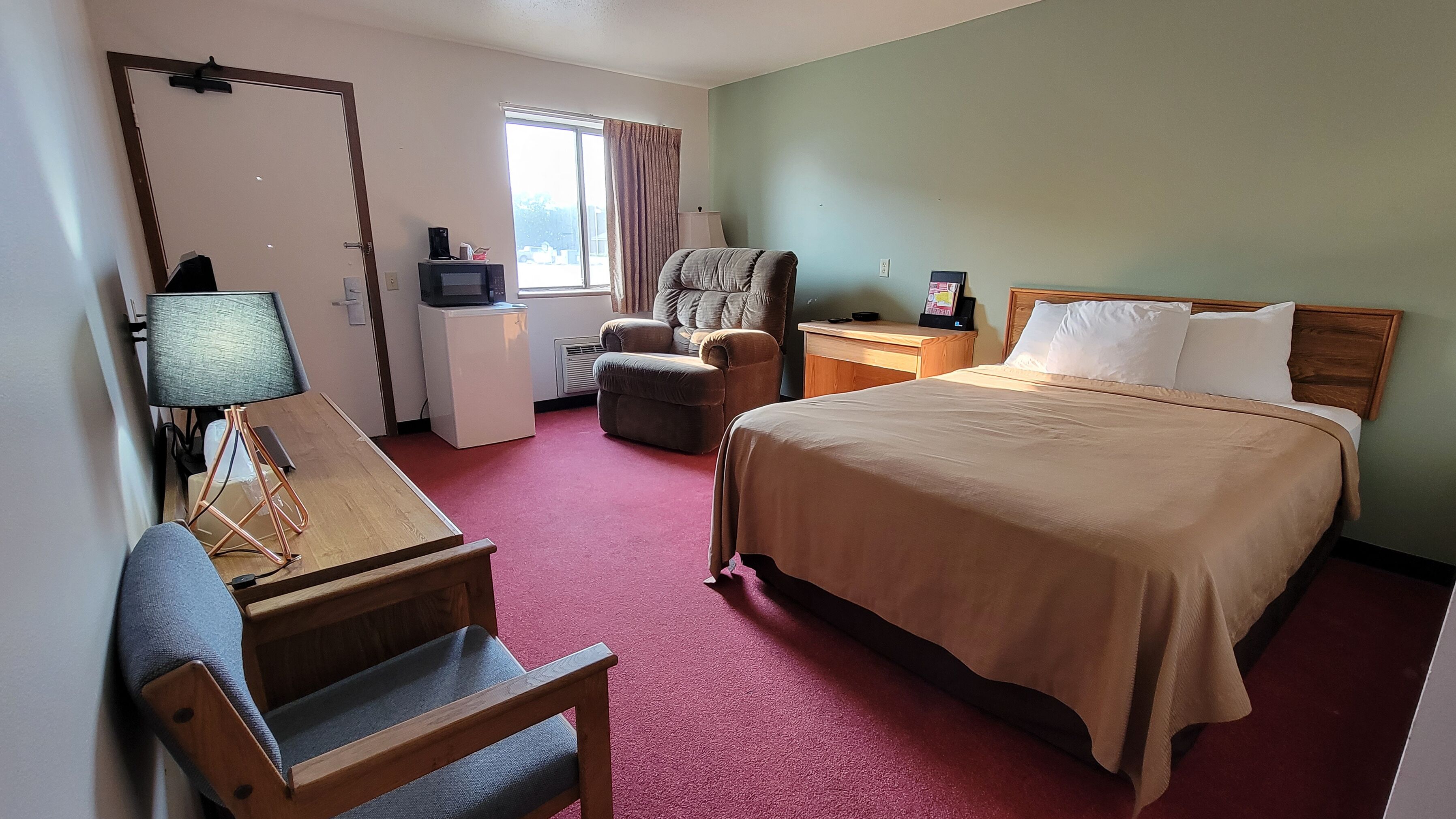 A hotel room with a bed, a desk with a lamp, a chair, a small table, and a television.