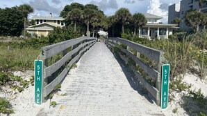 Ein Weg führt zu einem Strand, begleitet von einem Holzgeländer, Palmen und Gebäuden im Hintergrund.