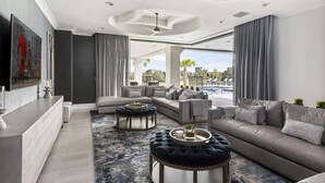A modern living room with a sectional sofa, ottomans, and a coffee table. There is a large window providing a view of a pool and palm trees.