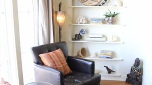 A living room with a black leather sofa, a patterned throw pillow, a wooden floor, a red patterned rug, and a wall-mounted shelf with decorative items. - Timeless Elegance ~ 1920's Sam Hughes Casita Mariposa ~ Walk to UA! Parking (Tucson)