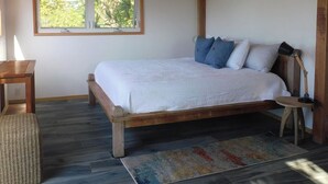 Un dormitorio con cama de madera, ventilador de techo, ventana con vista a vegetación, mesita de noche y alfombra.
