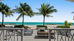 A wooden deck with lounge chairs and a table, overlooking a beach with palm trees and the ocean.