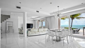 A spacious living area with a white sofa, dining table, and chairs, large windows with a view of the ocean, and a modern ceiling design.