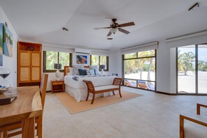 Une chambre spacieuse comprenant un lit, une chaise en bois, un bureau et une grande fenêtre donnant sur des palmiers et une plage.