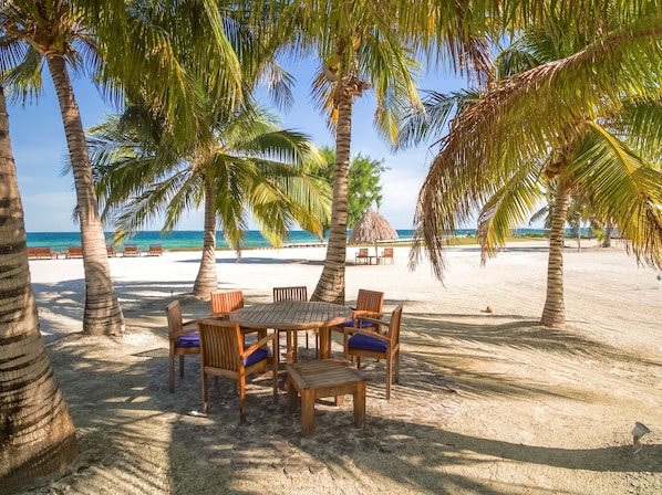 A beachside dining area with a round wooden table and chairs, surrounded by palm trees and a view of the ocean.