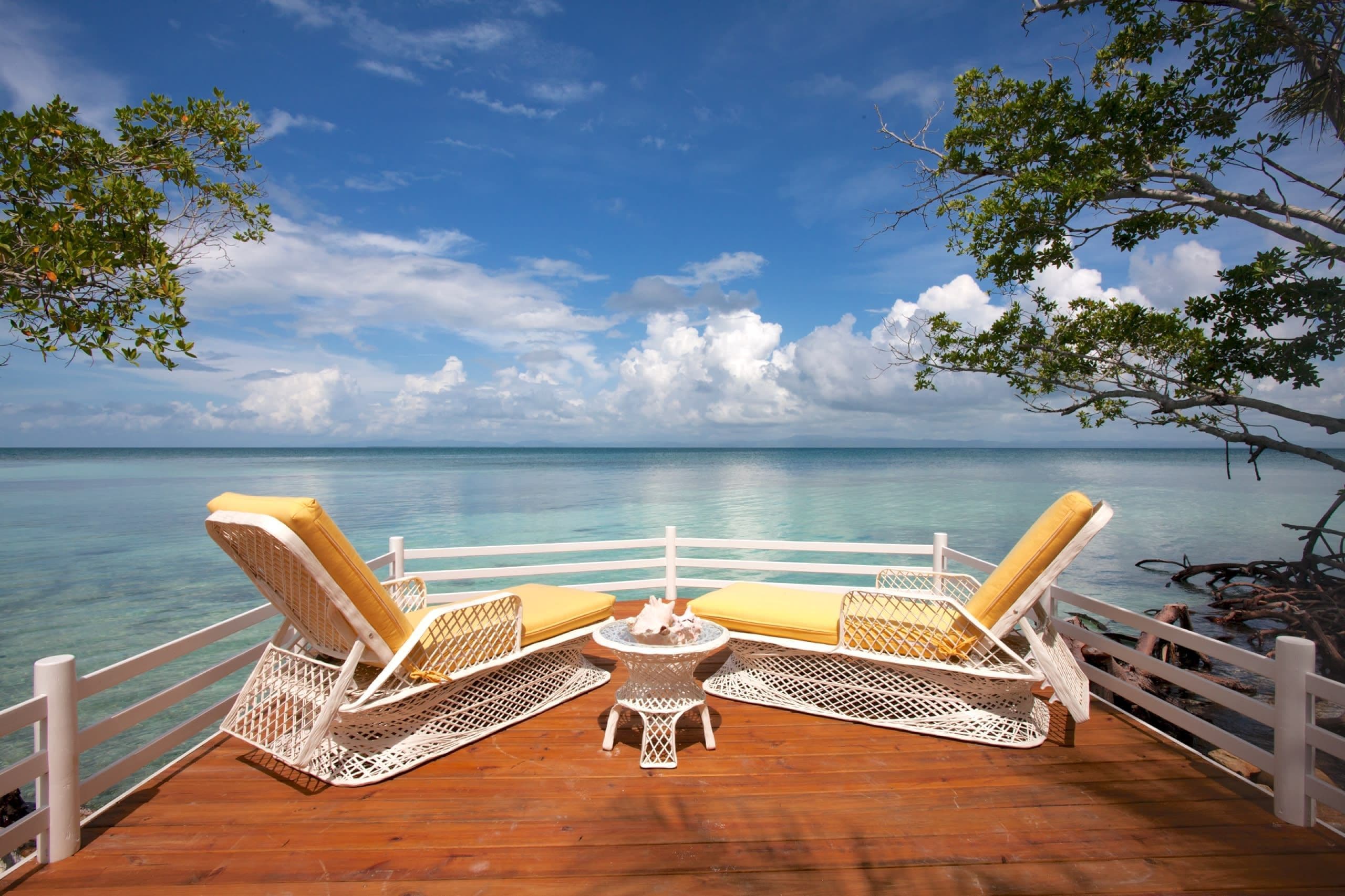 A wooden deck with two wicker lounge chairs and a small table, overlooking a clear blue sea.