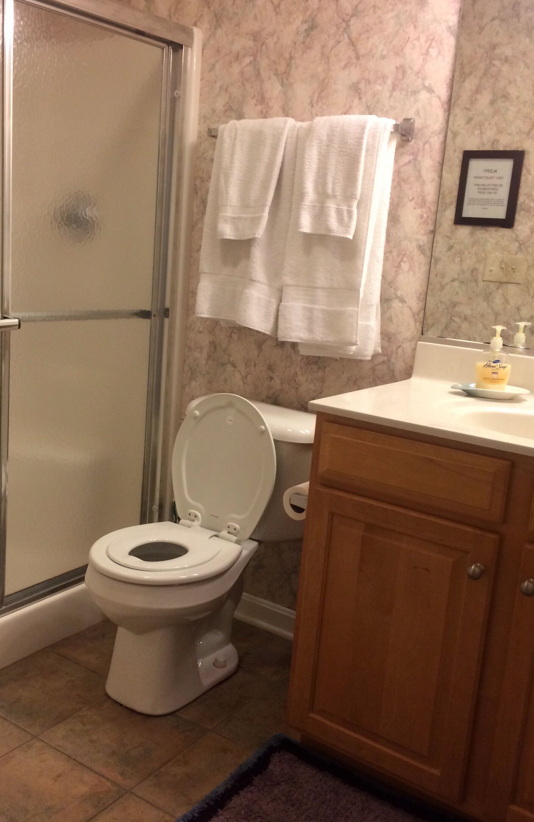 A bathroom with a toilet, a shower with a glass door, marble walls, and a wooden cabinet.