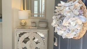 Une porte ornée d’un panier tressé rempli de coquillages, une console blanche avec une lampe et des objets décoratifs, et un miroir au-dessus de la console.