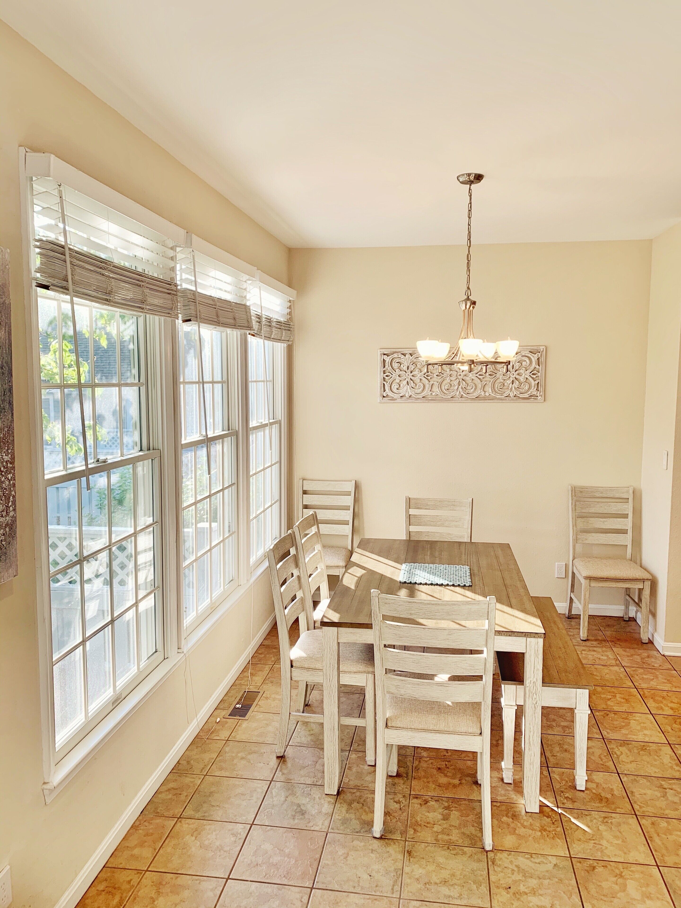 A dining area with a wooden table and chairs, large windows with blinds, a chandelier, and a decorative wall piece.