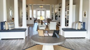 Un salon spacieux avec une table basse ronde, deux canapés blancs ornés de coussins bleus, un lustre et une grande fenêtre.