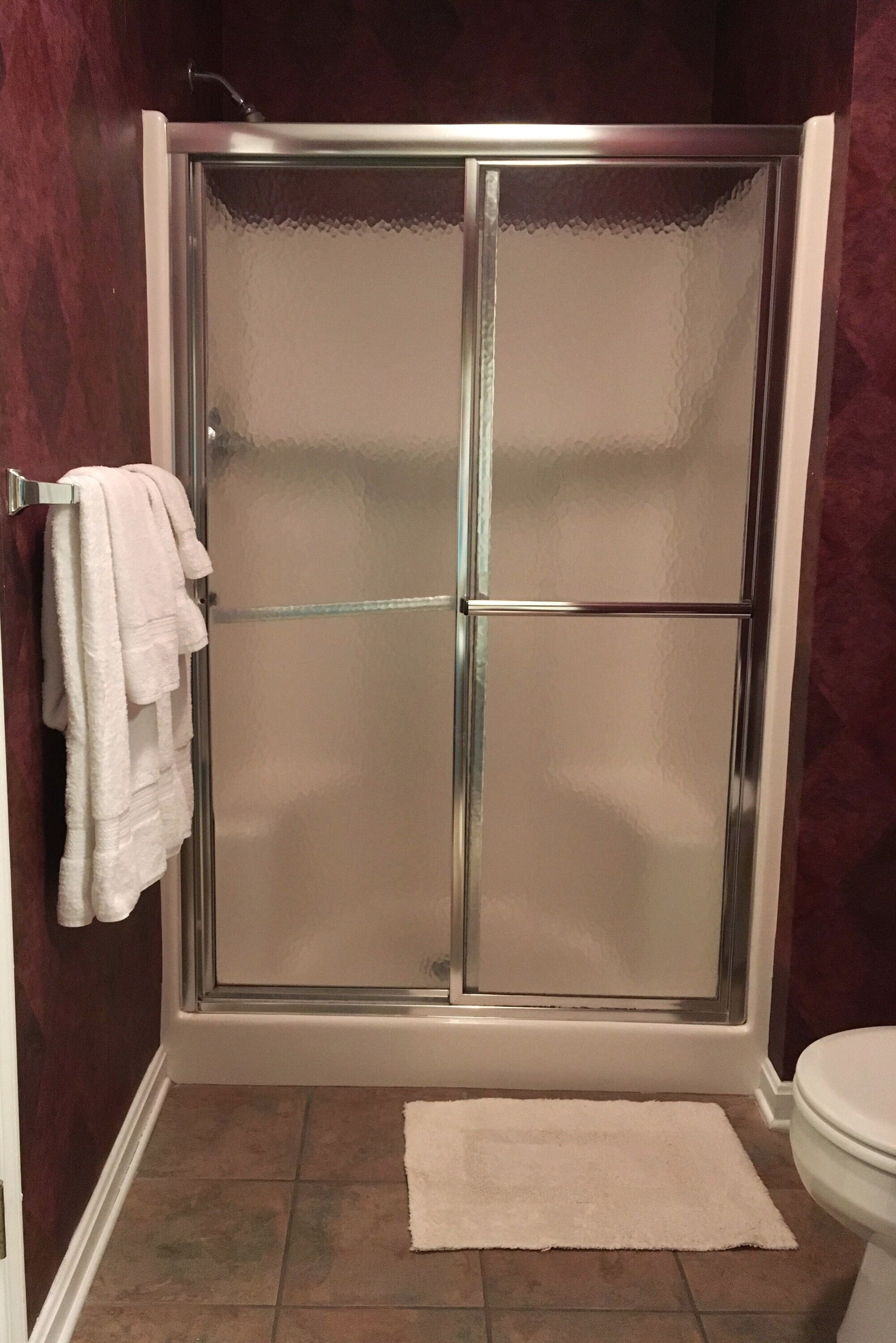 A shower with a glass door, a white towel hanging on a rack, and a white rug on the floor.
