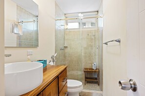 Un baño con un lavamanos grande de color blanco, un mueble de tocador de madera, un inodoro y una zona de ducha con mampara de cristal.