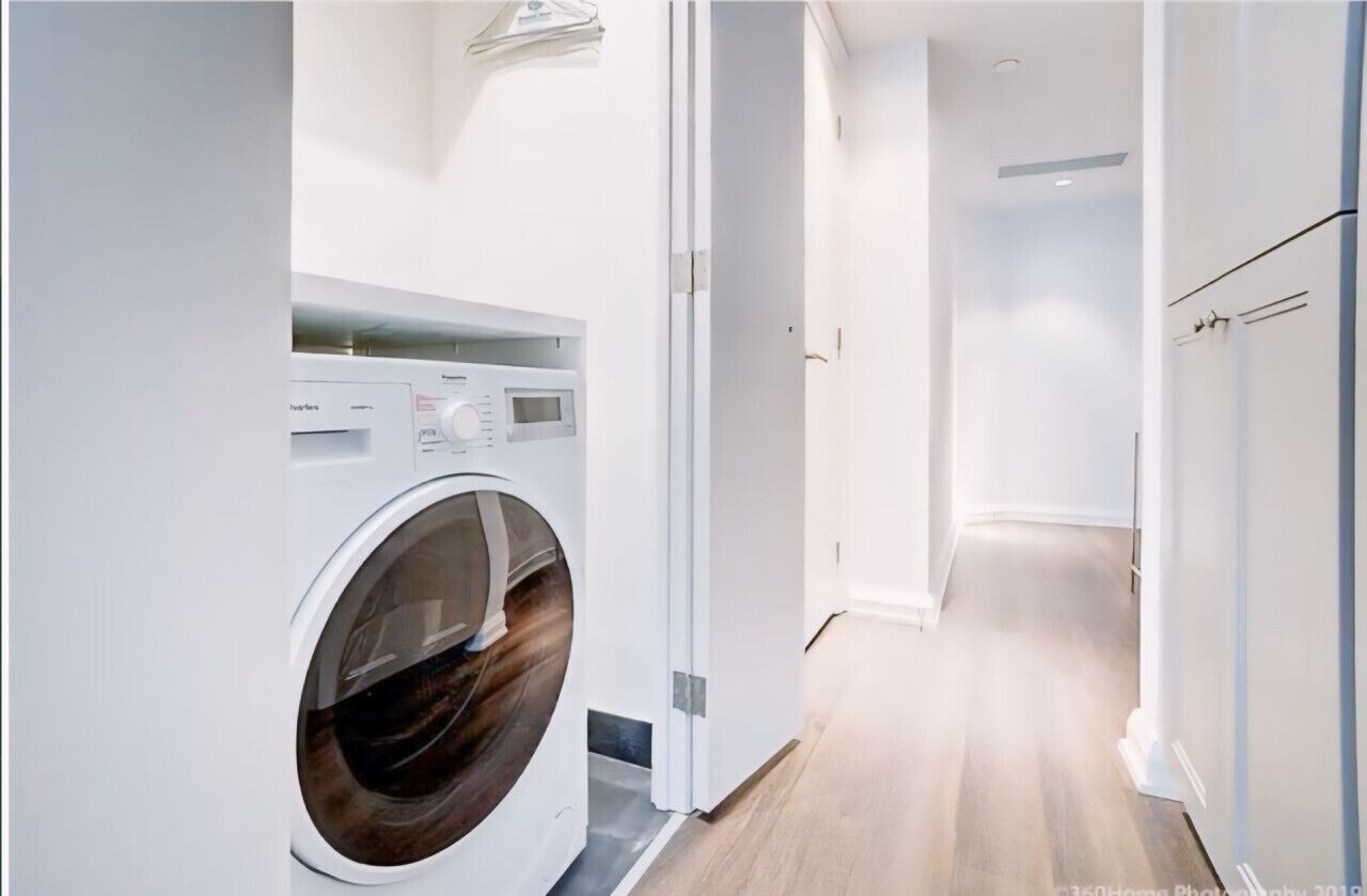 A modern laundry room with a front-loading washing machine, a hallway with wooden flooring, and white walls.