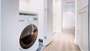 Une buanderie moderne équipée d’un lave-linge à chargement frontal, un couloir avec un plancher en bois et des murs blancs.