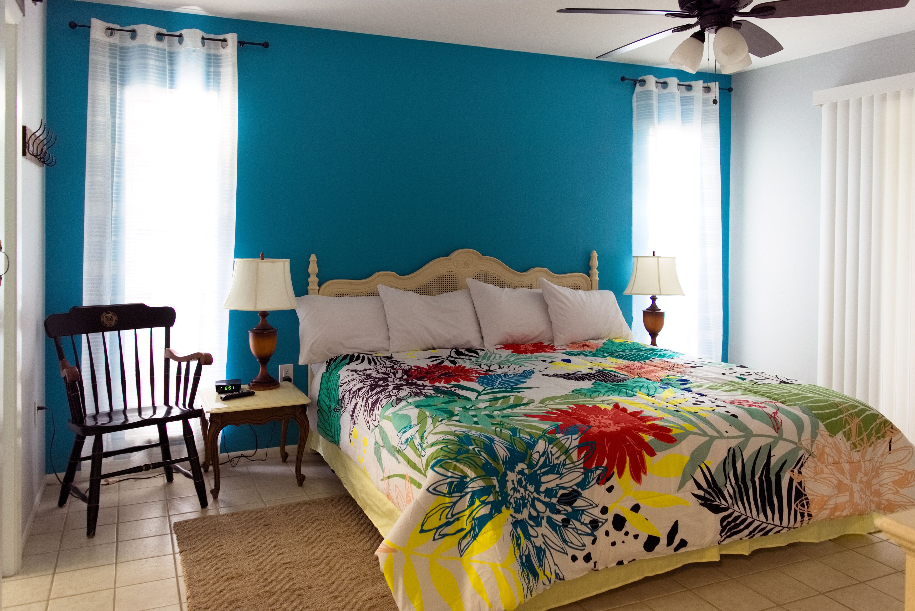 A bedroom with a blue wall, a wooden bed with a floral bedspread, two bedside lamps, a wooden chair, and a small table with a lamp.