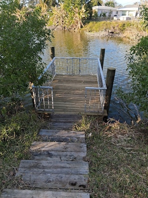 Une jetée en bois menant à une étendue d’eau, entourée de verdure et avec une zone résidentielle en arrière-plan.
