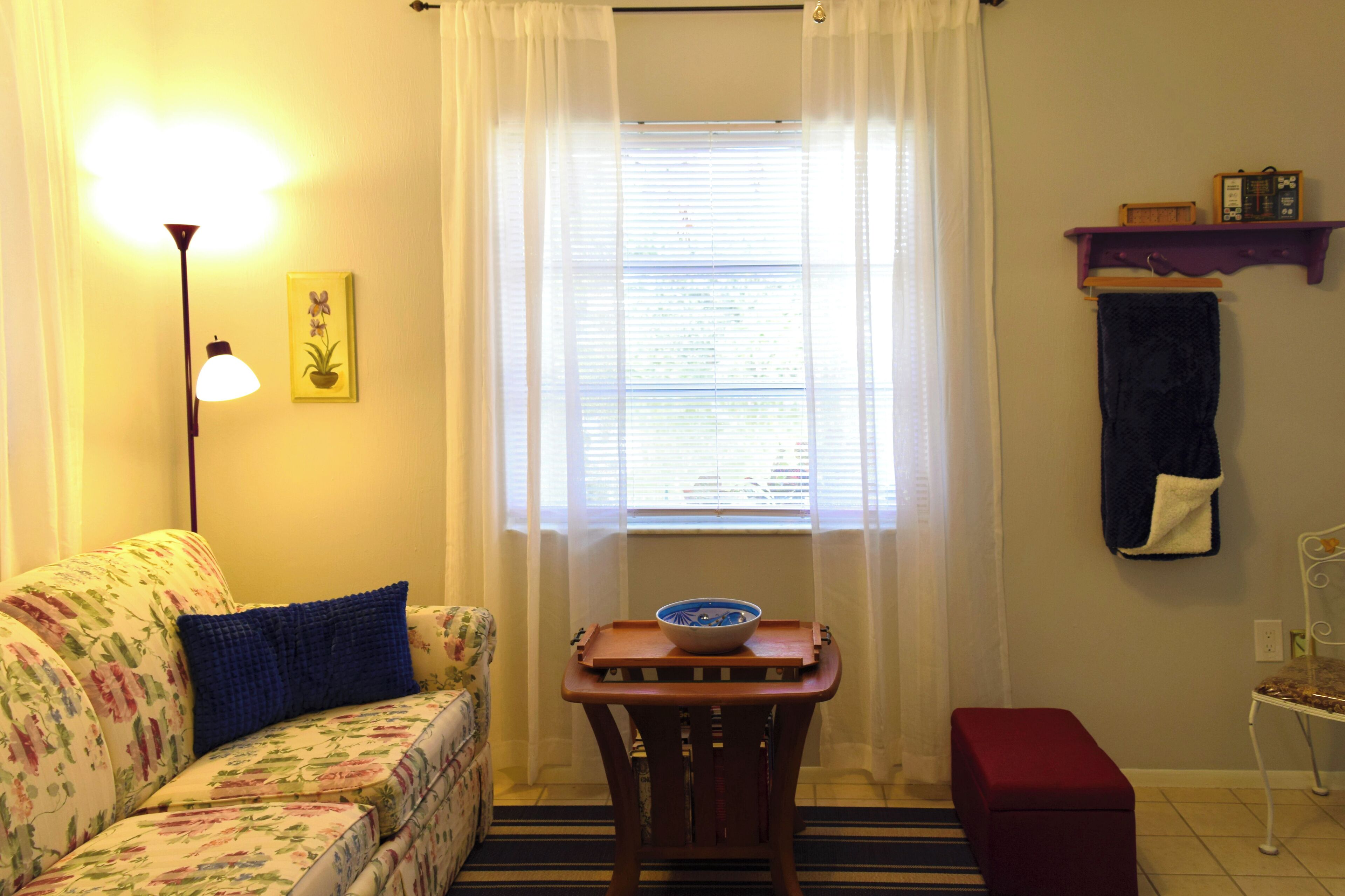 A living room with a floral sofa, a small wooden table with a bowl, a standing lamp, a window with curtains, a shelf with a clock, and a red bench.