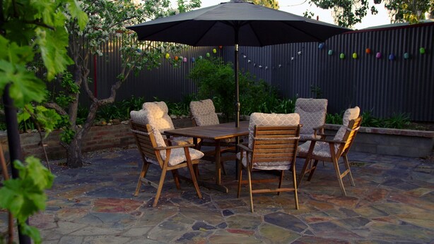 Patio esterno con pavimento in pietra, tavolo in legno e sei sedie con sedili imbottiti.