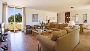 A spacious living room with a large sofa, a coffee table, and a dining area. There is a sliding glass door leading to a balcony with a view of greenery. - Luxury modern stone clad Villa with swimming pool and mountain views (POLLENSA)