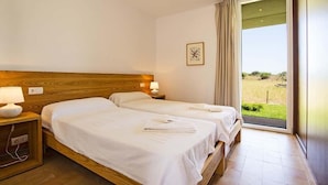 A hotel room with two beds, a wooden headboard, a nightstand with a lamp, and a view of a grassy area through a large window. - Luxury modern stone clad Villa with swimming pool and mountain views (POLLENSA)