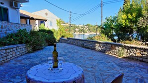A stone patio with a table set for two, overlooking a body of water and a hillside. - Angelina: Charming studio, terrace views, steps from Kalami beach (Kalami)
