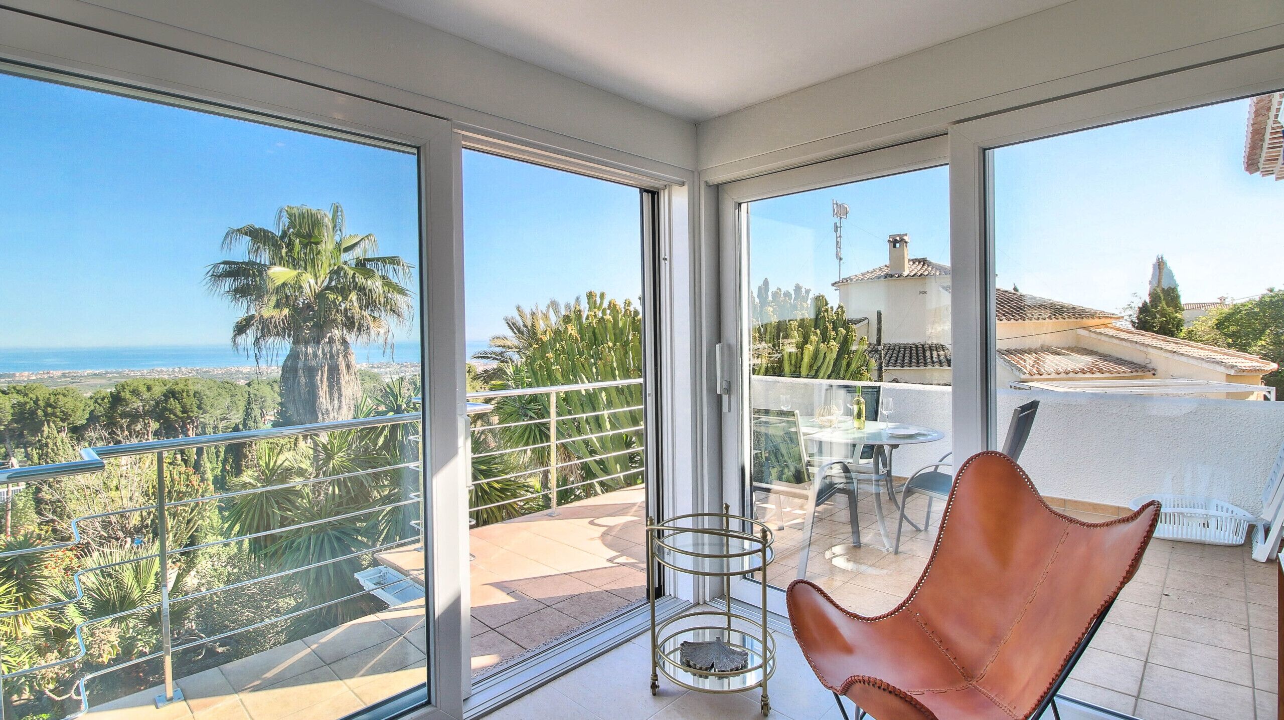Een balkon met een glazen deur, een tafel met stoelen en uitzicht op zee en palmbomen.
