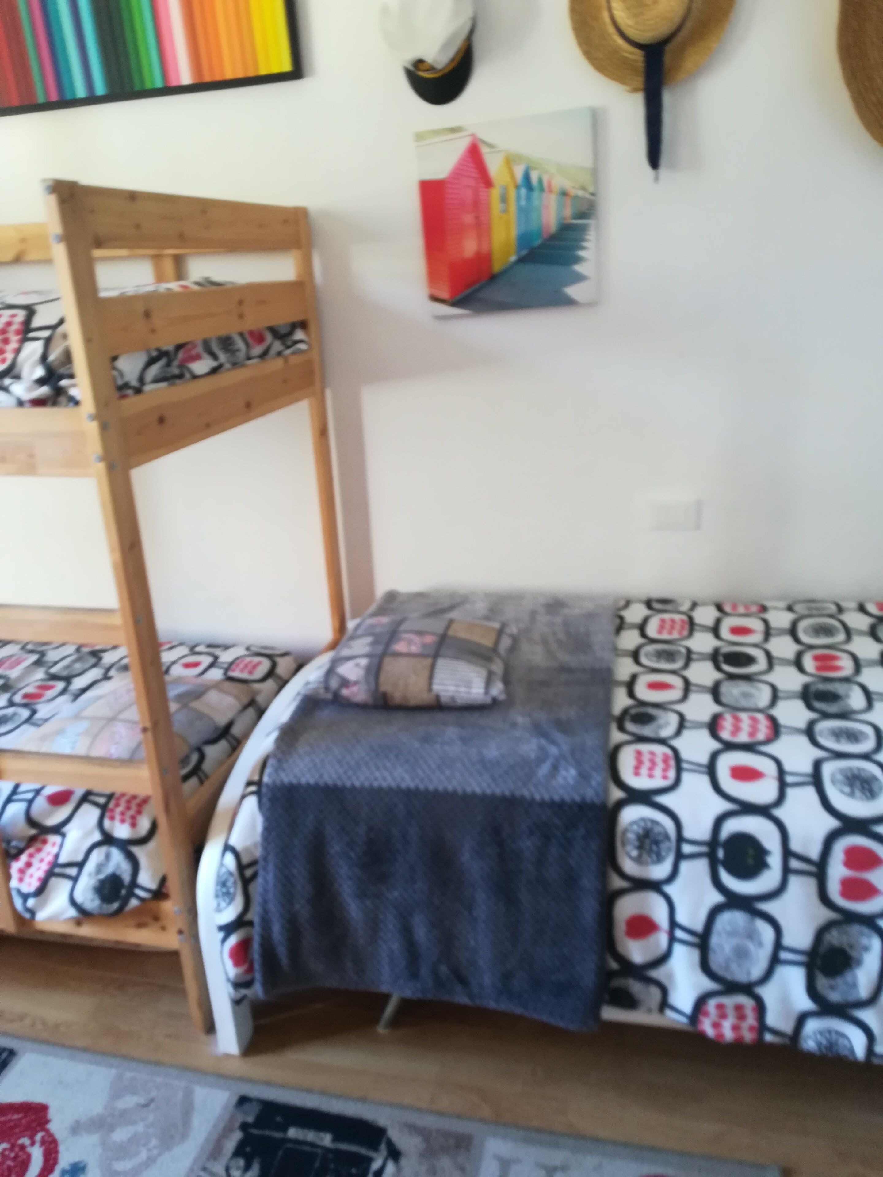 A bunk bed with a patterned bedspread and pillows, a wooden chair with a patterned cushion, and a wall with a colorful painting and a hat hanging on the wall.