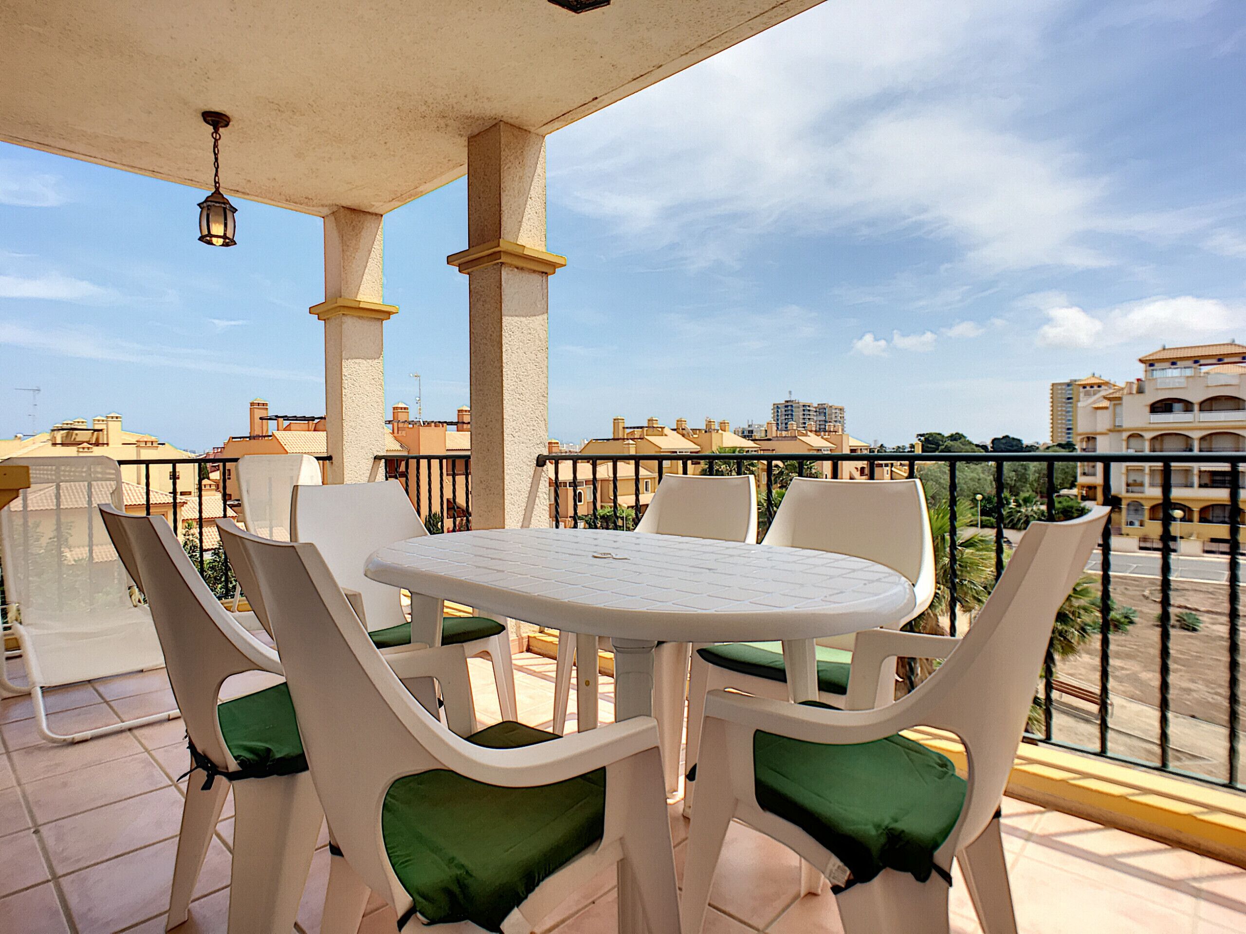 Eine Terrasse mit rundem Tisch und Stühlen, einer hängenden Lampe und Blick auf Gebäude im Hintergrund.