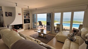 Ein geräumiges Wohnzimmer mit Kamin, einem großen Fenster mit Meerblick, einem Pool und einem Balkon mit Gartenmöbeln.
