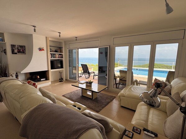 A spacious living room with a fireplace, a large window with a view of the ocean, a pool, and a balcony with outdoor furniture.