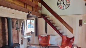A cozy cabin interior with a wooden ceiling, a large woven light fixture, a rustic wooden table, a patterned rug, and a stone fireplace.