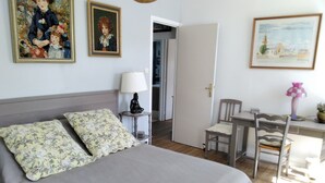 Een slaapkamer met een bed, twee kussens, een nachtkastje met een lamp, een stoel, een bureau met een lamp en twee ingelijste foto’s aan de muur.