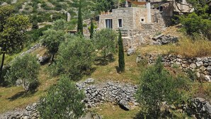 Un bâtiment en pierre surmonté d’un toit de tuiles rouges, entouré d’oliviers et délimité par un mur de pierre.