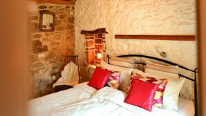 Une chambre avec un mur en pierre, des poutres en bois au plafond, un lit avec une literie blanche et des oreillers rouges, un luminaire suspendu et une petite fenêtre.