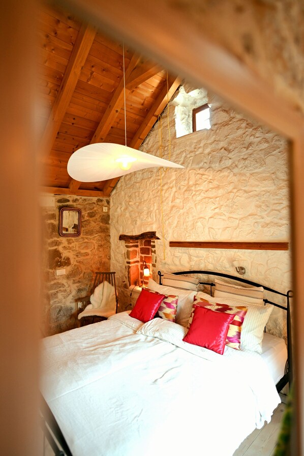A bedroom with a stone wall, wooden ceiling beams, a bed with white bedding and red pillows, a hanging light fixture, and a small window. - Private Boutique Retreat Surrounded by Pure Nature (Hvar)
