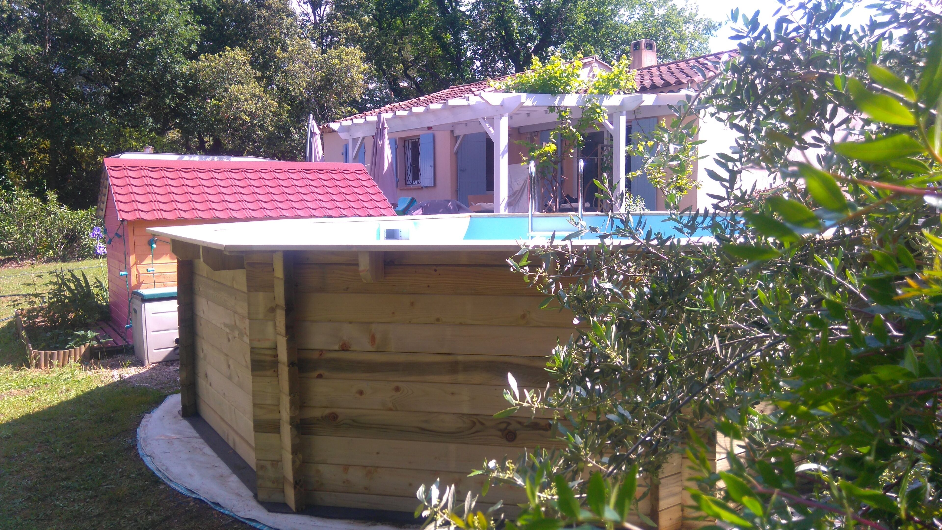 Una estructura de madera con piscina, rodeada de vegetación y una casa con techo rojo.