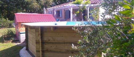 Una estructura de madera con piscina, rodeada de vegetación y una casa con techo rojo.