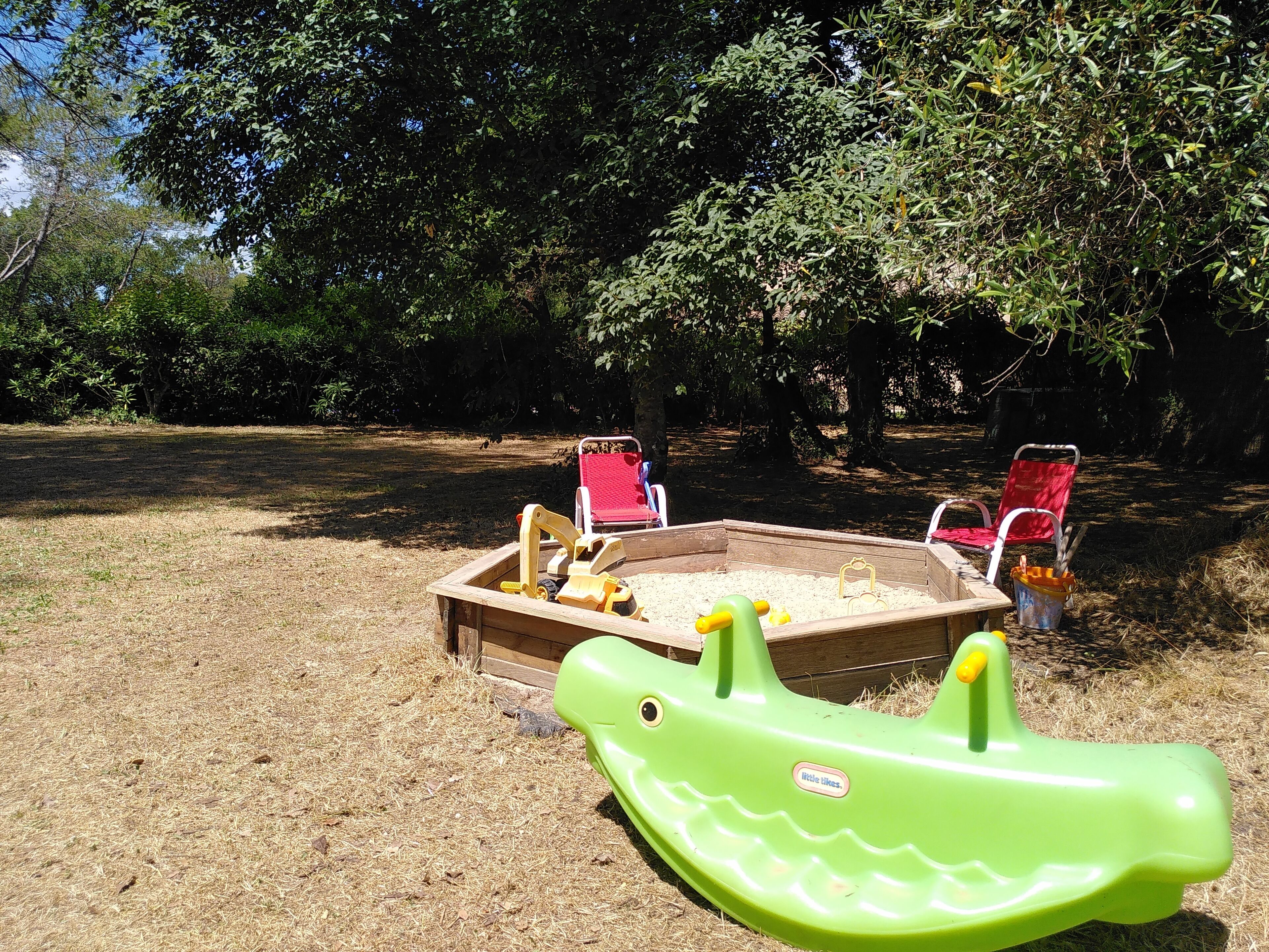 A sandbox with a green toy dinosaur, two red chairs, and a wooden play structure in a grassy outdoor area.