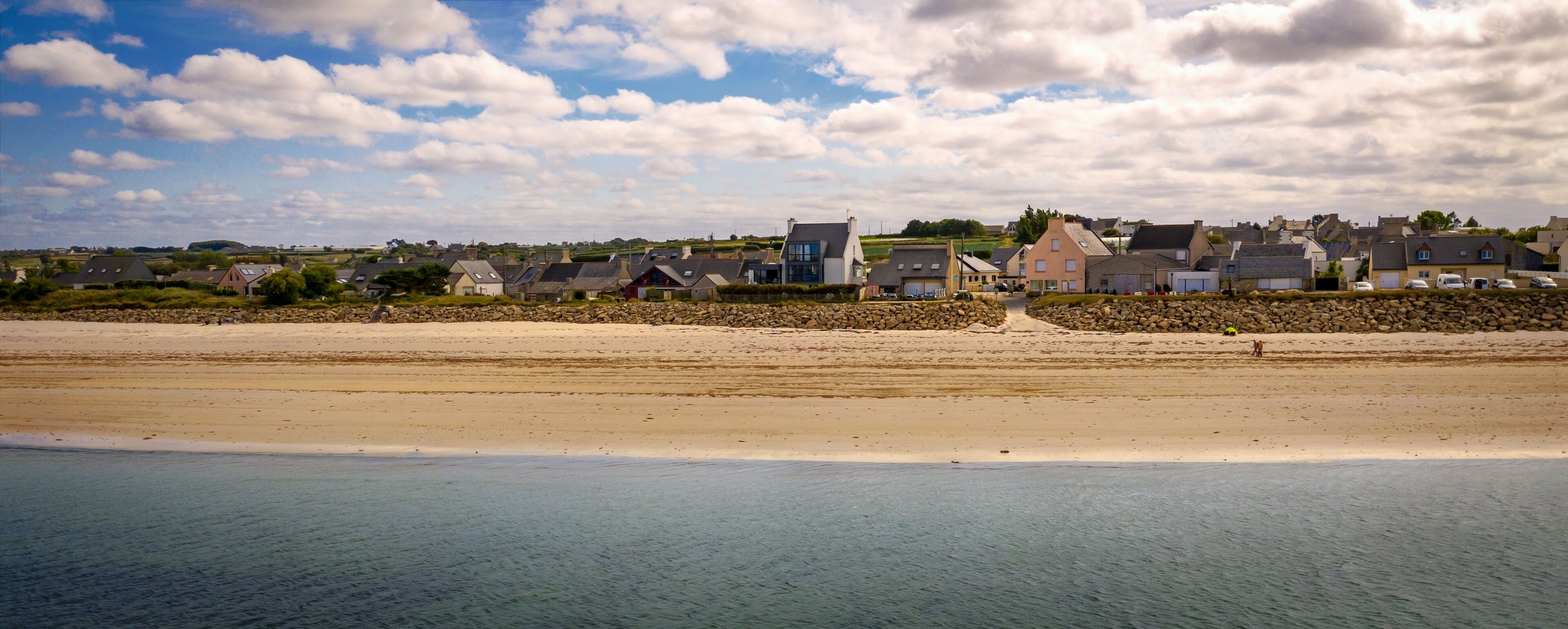 A coastal town with houses along the beach, a stone wall, and a clear sky.