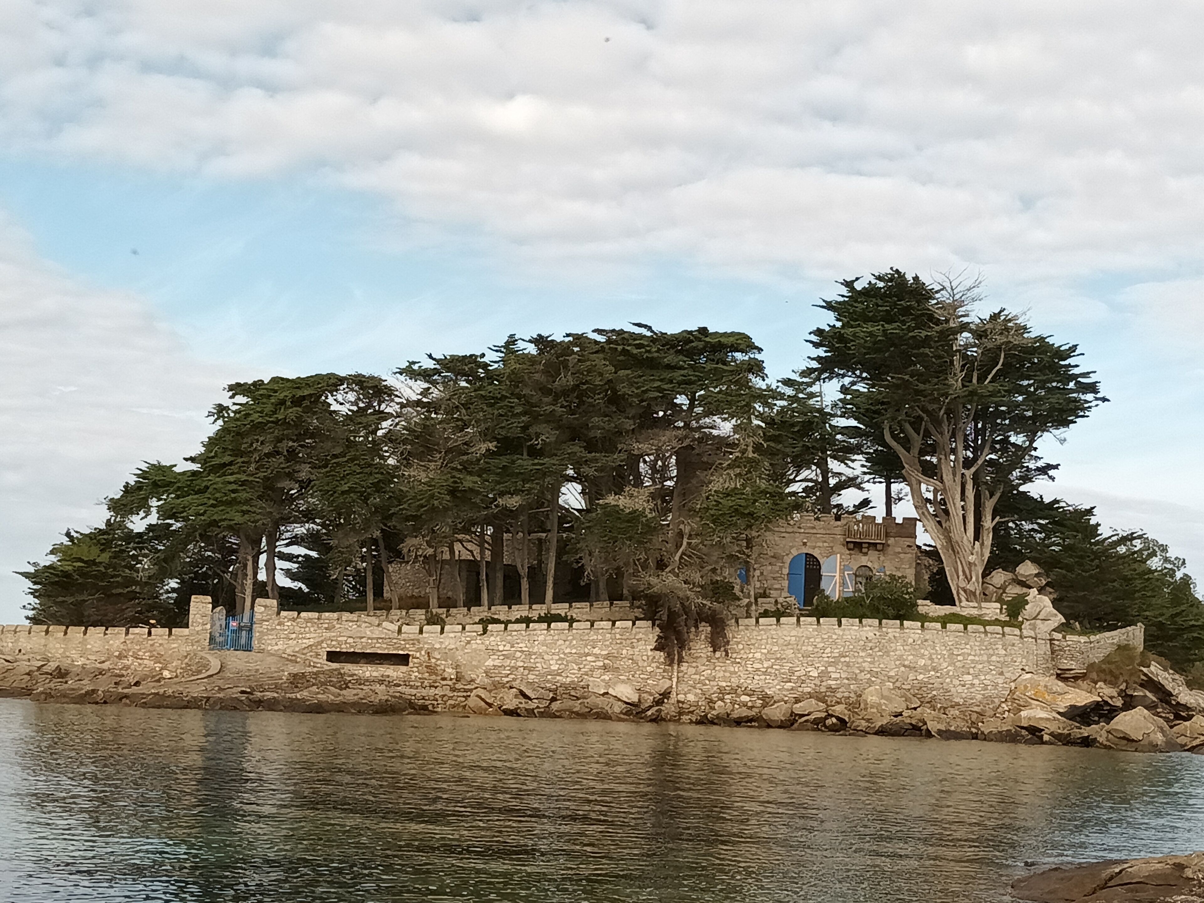 Un mur de pierre avec une porte bleue, des arbres et une étendue d’eau.