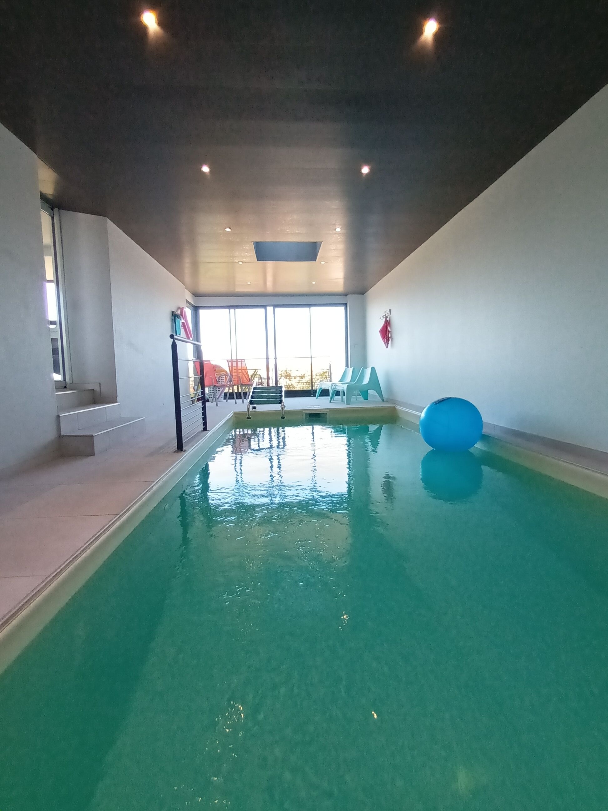 Une piscine intérieure à l’eau claire, avec des marches permettant d’y accéder, et une vue sur l’extérieur grâce à de grandes fenêtres.
