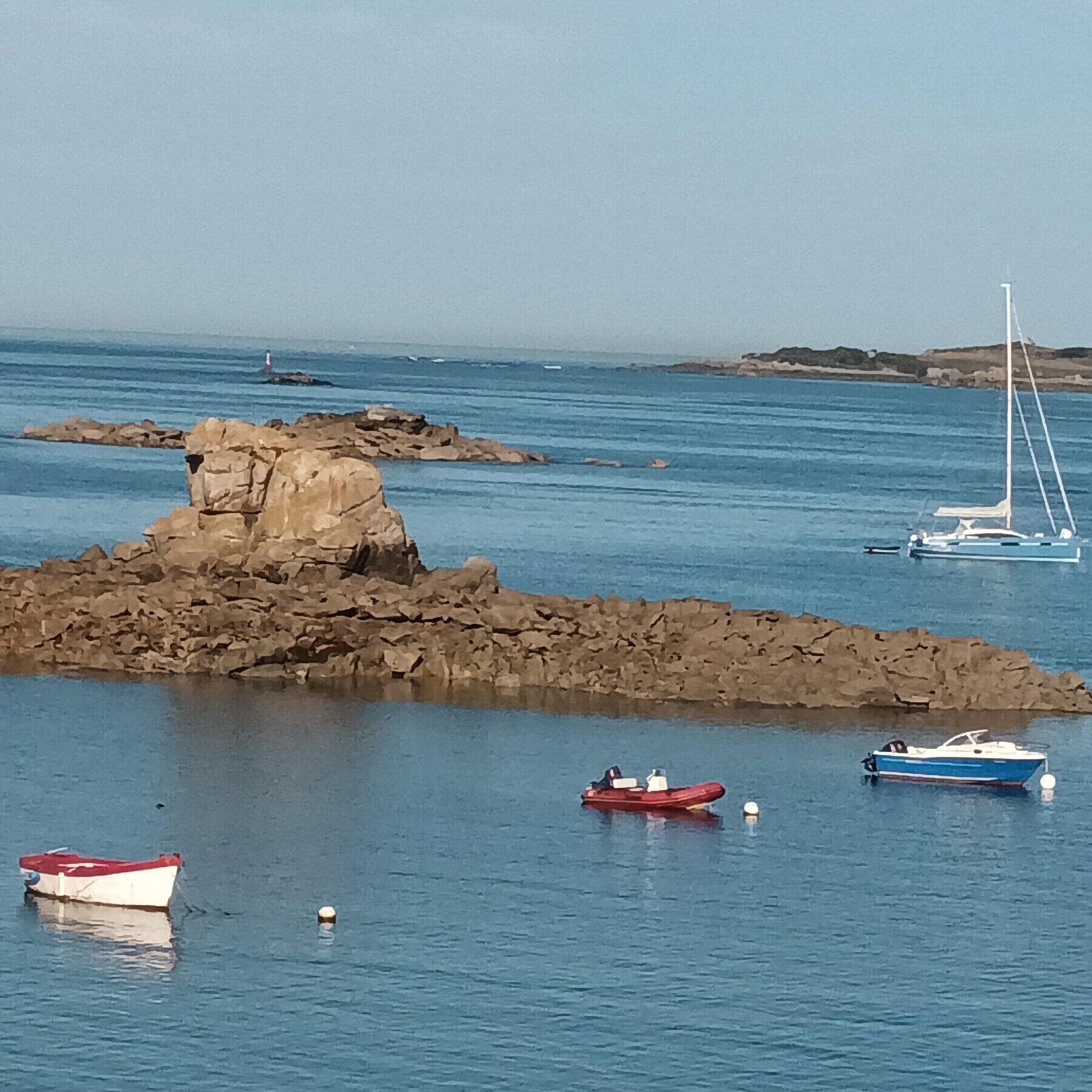 A coastal scene with rocky outcrops, calm water, and several boats including a sailboat and motorboats.