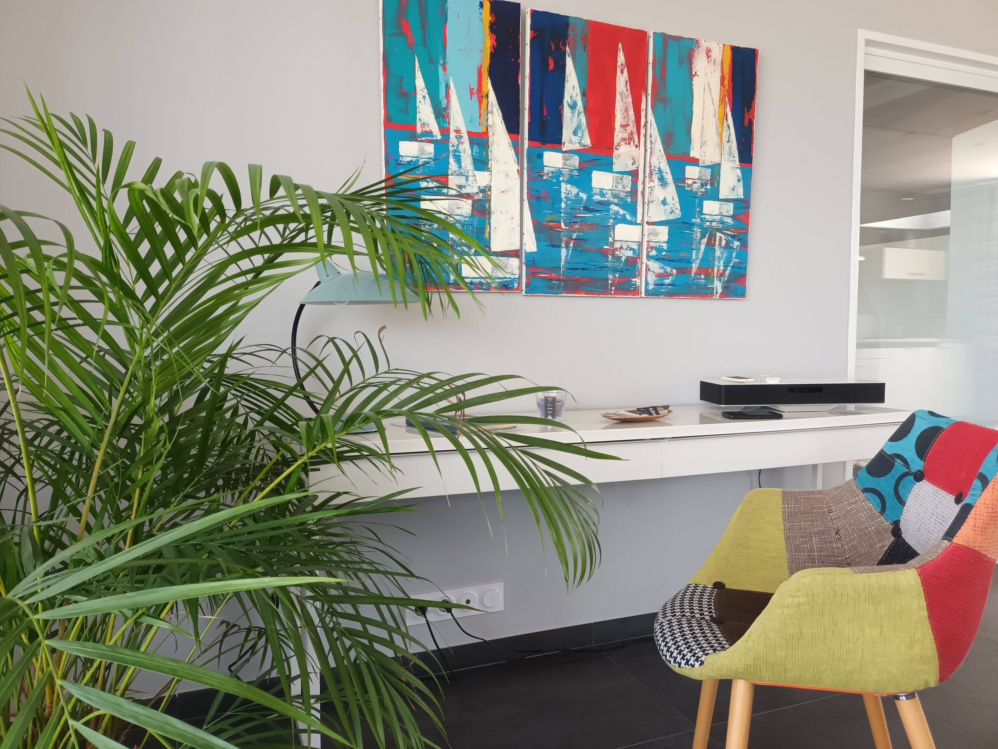 A modern office space with a white desk, a colorful chair, a large potted plant, and a vibrant abstract painting on the wall.